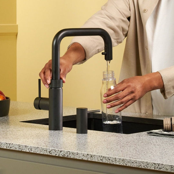 Person using the Quooker Flex Square black boiling water tap to fill a glass bottle in a modern kitchen, available at Spoelbakhuis.