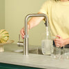 Person filling a glass carafe using the Quooker Flex Square stainless steel boiling water tap in a modern kitchen, available at Spoelbakhuis.